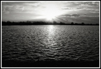 A heavy contrasted black and white image of the sun going down over a large lake
