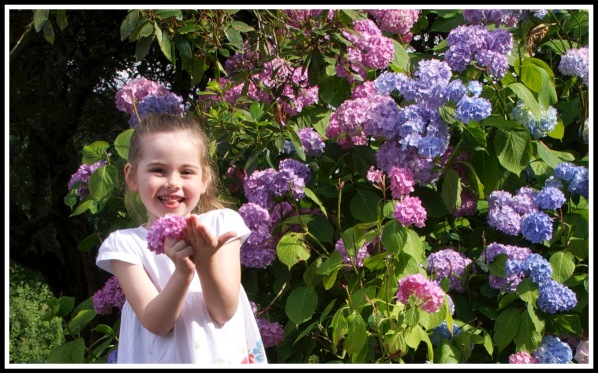 Ella smiling with a bunch of flower, stood next to a vividly coloured bush with lots of flowers
